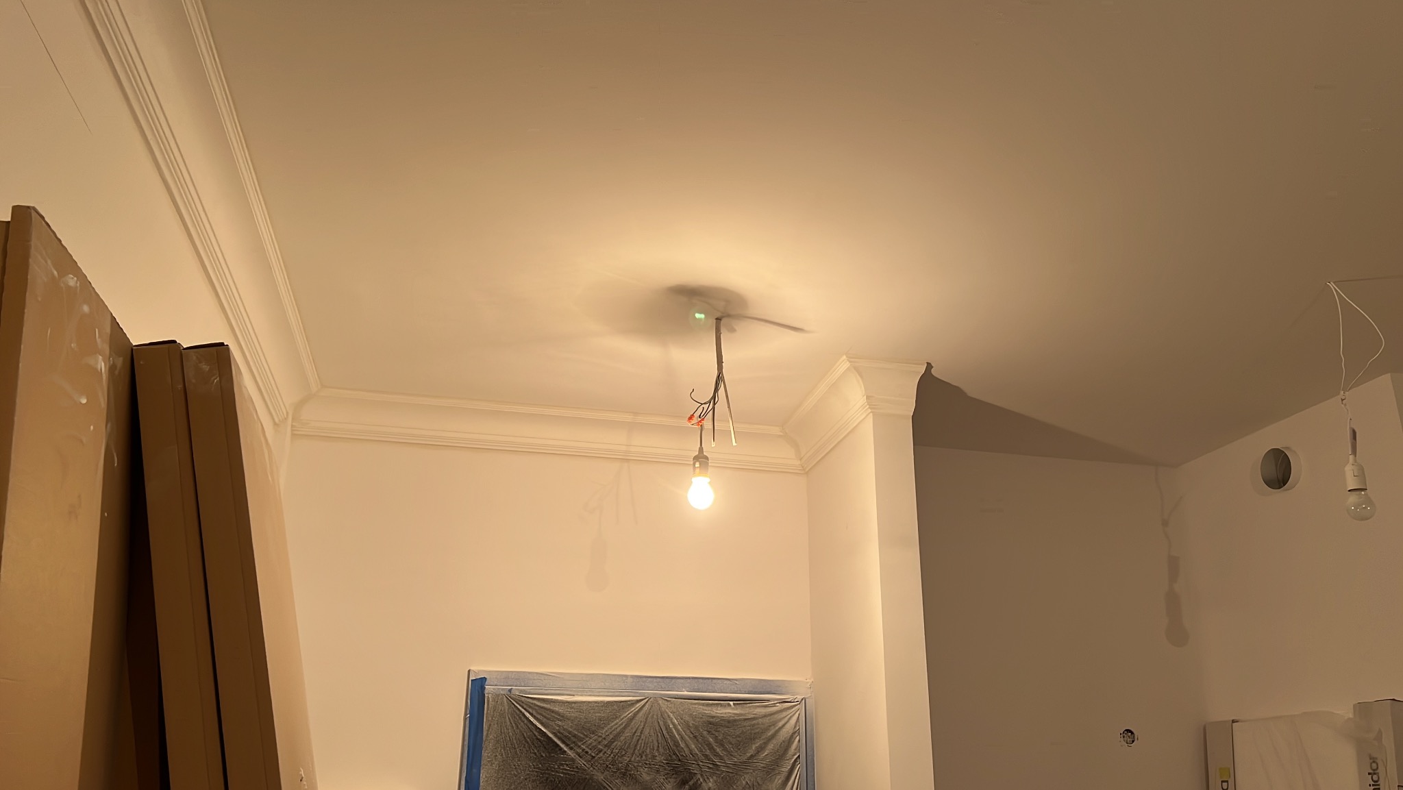 Wnętrze w trakcie prac wykończeniowych: pomalowane ściany, listwy dekoracyjne pod sufitem, tymczasowe oświetlenie. Okno zabezpieczone folią.