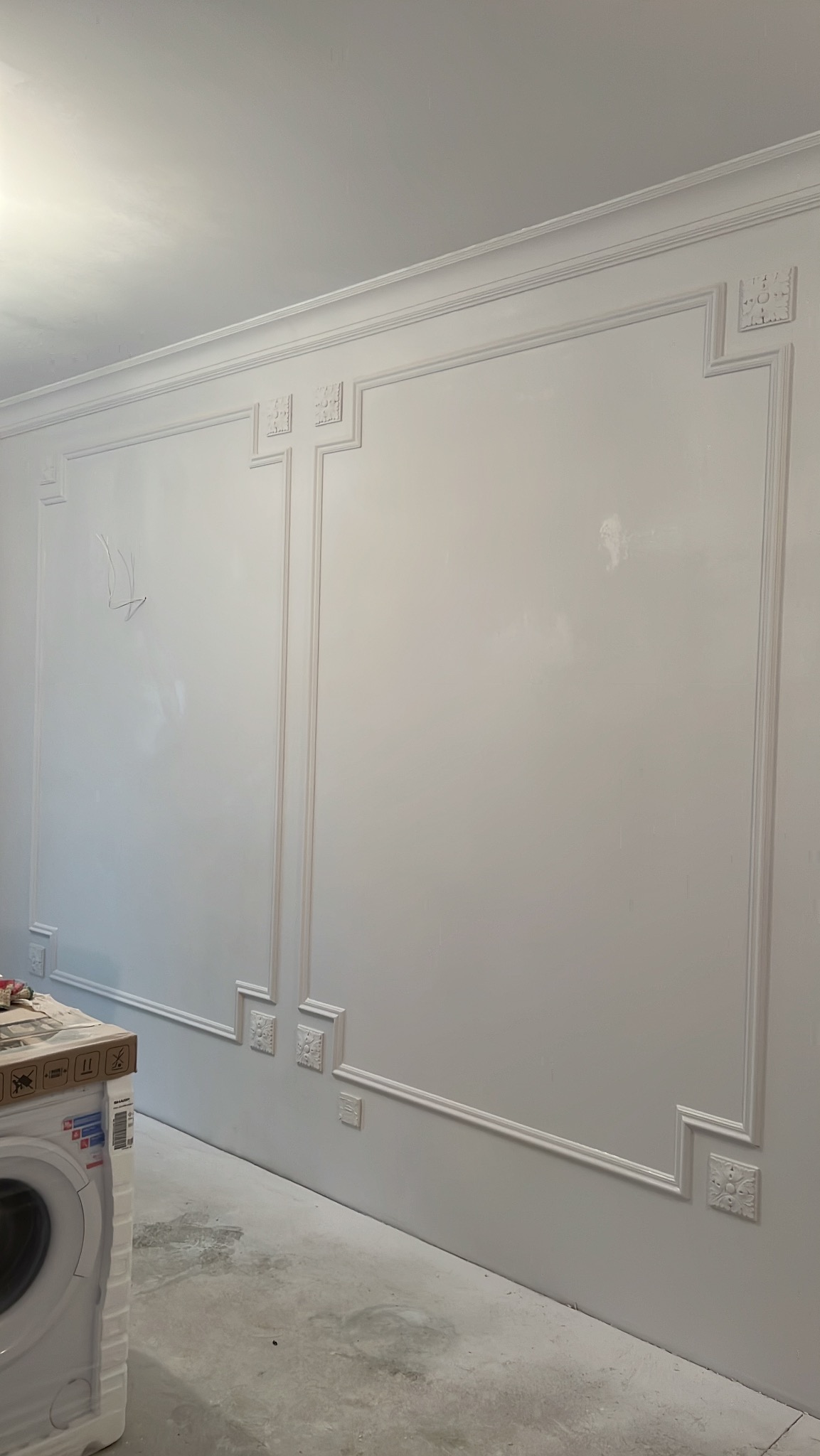 Biała ściana z dekoracyjnymi listwami i ornamentami w stylu klasycznym. Widoczny fragment pralki w folii ochronnej. Świeżo po malowaniu.