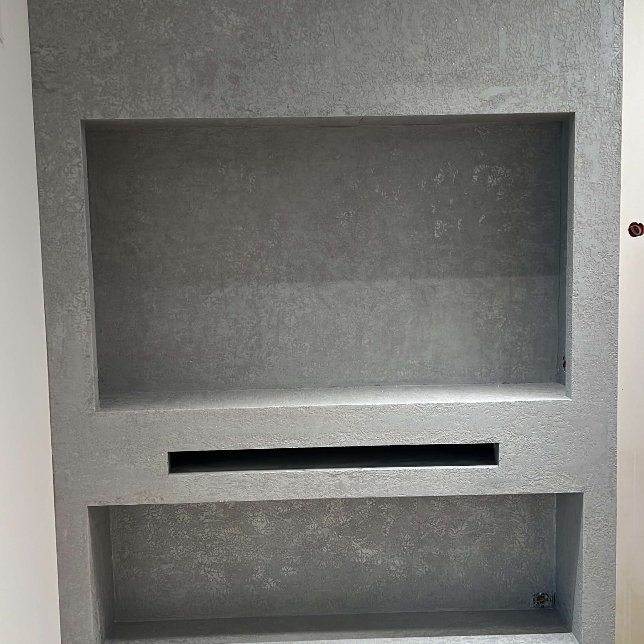 Szara ściana z wnękami o nieregularnej fakturze, wykończona tynkiem dekoracyjnym. Widoczne dwie półki i wąski otwór wentylacyjny. Świeżo położony tynk.