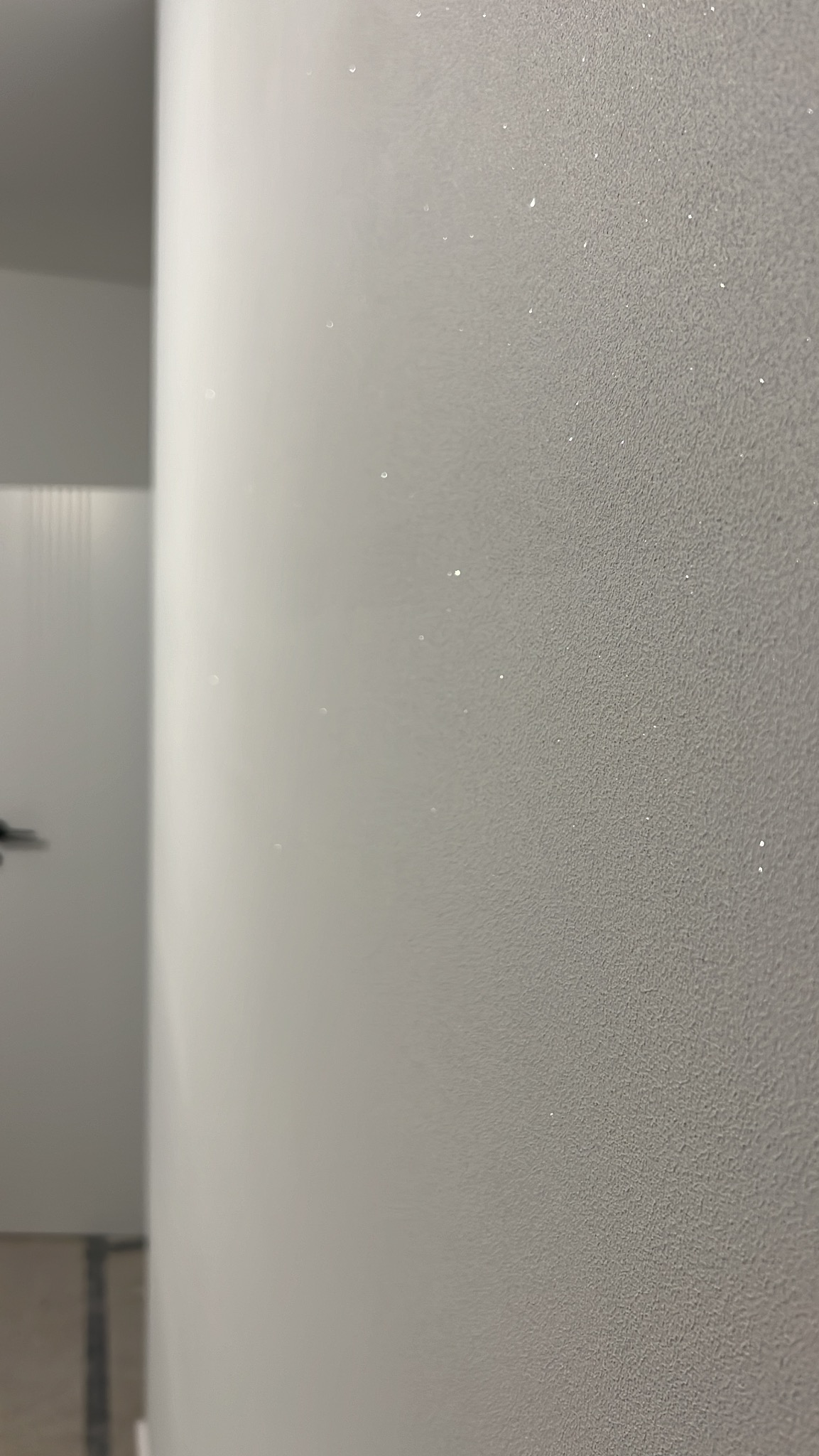 Szara ściana z delikatną, połyskującą fakturą, oświetlona z lewej strony, z widocznym fragmentem drzwi w tle. Minimalistyczne wykończenie wnętrza.