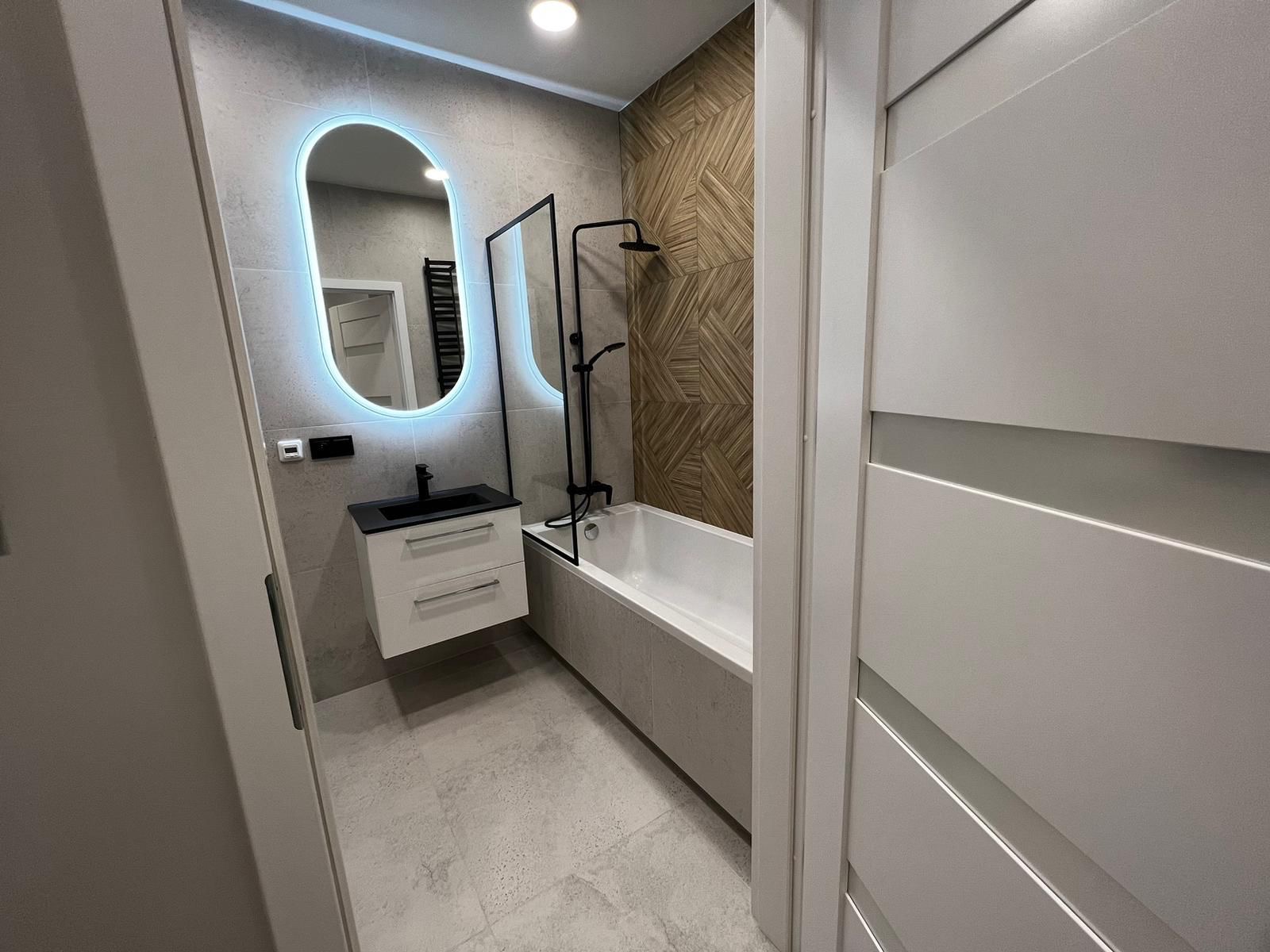 Nowoczesna łazienka po remoncie: owalne lustro LED, czarna armatura, płytki imitujące drewno i minimalistyczna zabudowa wanny. Widok zza uchylonych drzwi.