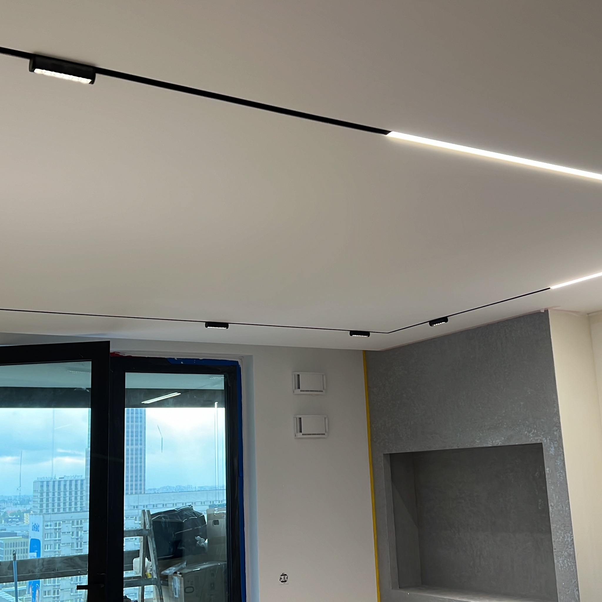 Nowoczesne oświetlenie sufitowe: czarne szyny z punktowymi i liniowymi lampami LED w jasnym wnętrzu z widokiem na miasto przez okno.