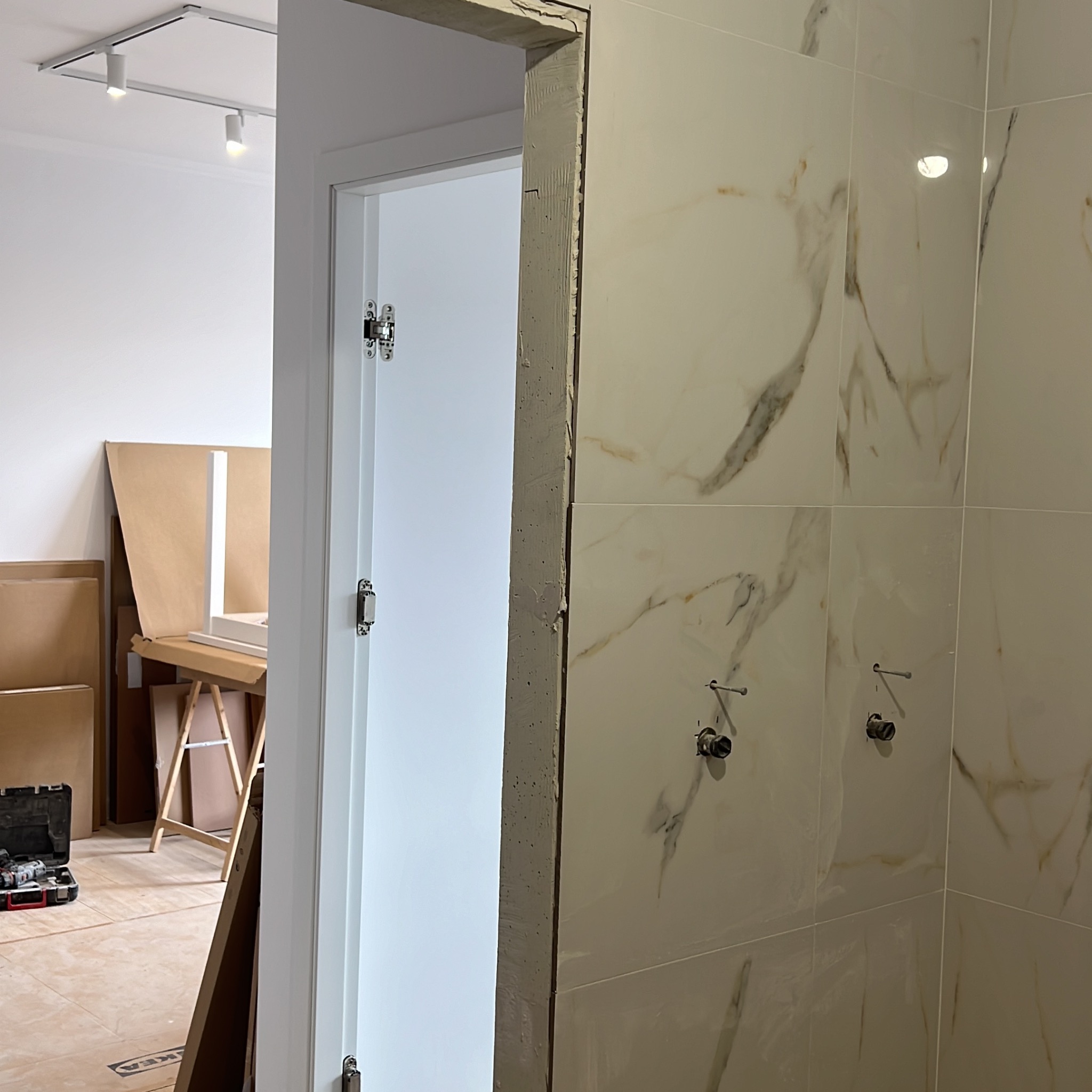Montaż drzwi i wykończenie łazienki z białymi płytkami imitującymi marmur. Widoczna surowa ościeżnica i narzędzia w tle, co sugeruje etap prac wykończeniowych.