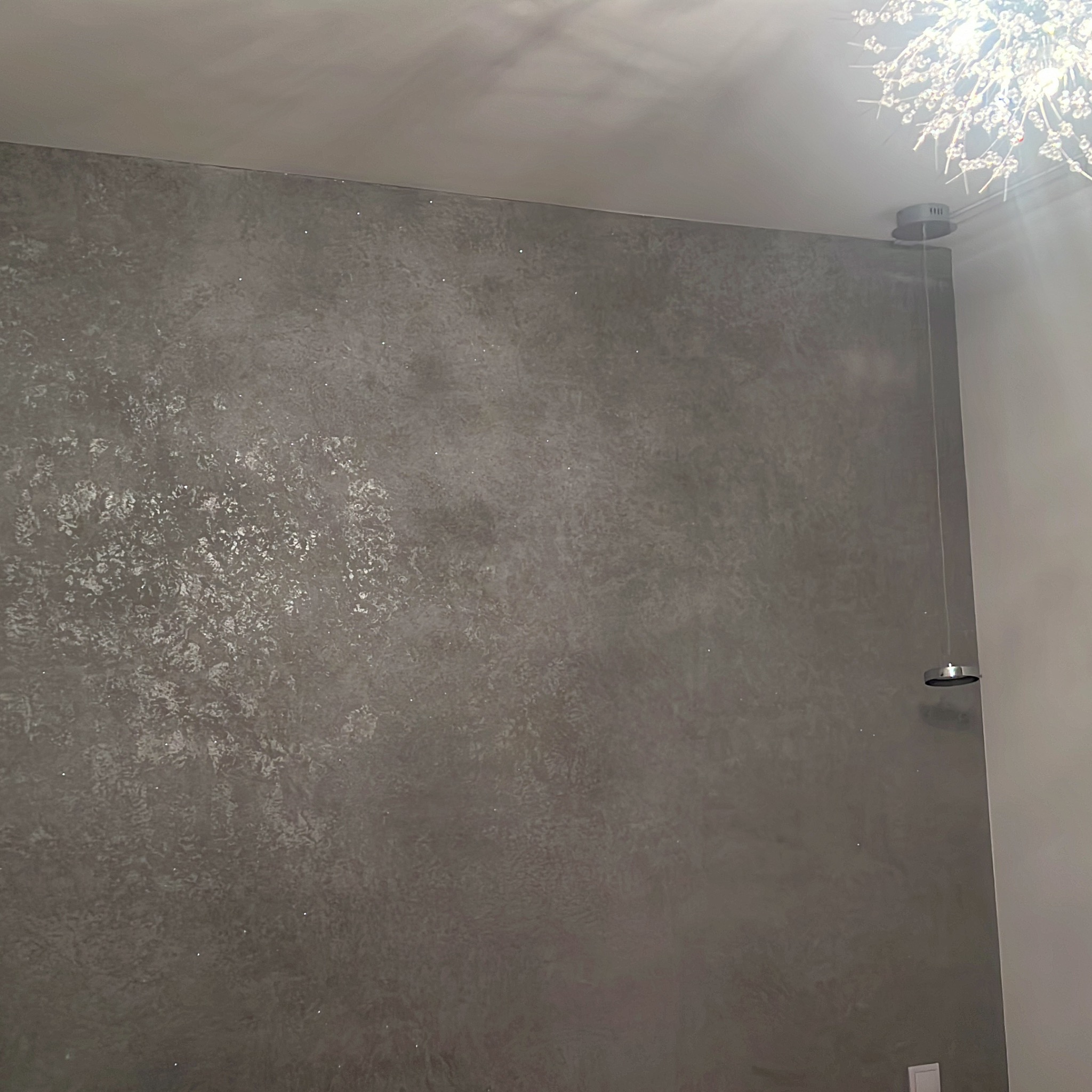 Ściana z dekoracyjnym, szarym tynkiem strukturalnym, oświetlona przez nowoczesną lampę sufitową. Widoczny fragment gładkiej, białej ściany obok.