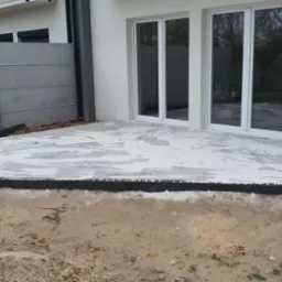 Układanie szarych płyt tarasowych przy białym domu z dużymi oknami. Widoczny fragment betonowego ogrodzenia i obrzeże z ciemnej kostki. Etap prac przed fugowaniem.