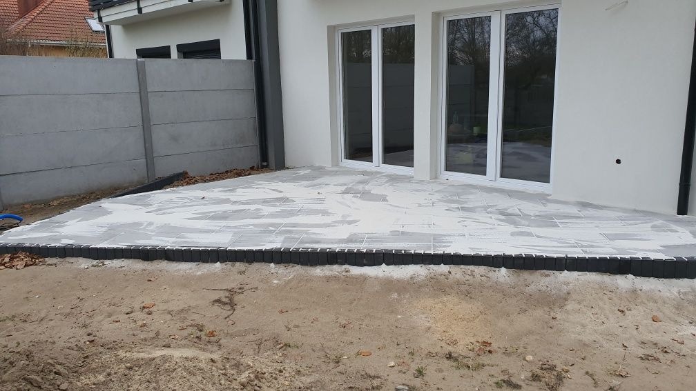Układanie szarych płyt tarasowych przy białym domu z dużymi oknami. Widoczny fragment betonowego ogrodzenia i obrzeże z ciemnej kostki. Etap prac przed fugowaniem.