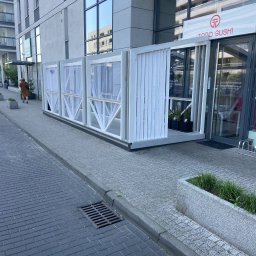 CARPENTER HUBERT GRUDZIŃSKI - Biała, drewniana konstrukcja z zasłonami przed wejściem do restauracji Todo Sushi. Konstrukcja osłania stoliki na zewnątrz. Widoczny fragment chodnika i elewacji budynku.