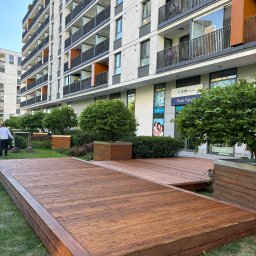 CARPENTER HUBERT GRUDZIŃSKI - Nowoczesny budynek z balkonami i drewnianym tarasem na zielonym trawniku. Uwagę przyciąga starannie wykonany taras z drewna o ciepłym odcieniu.