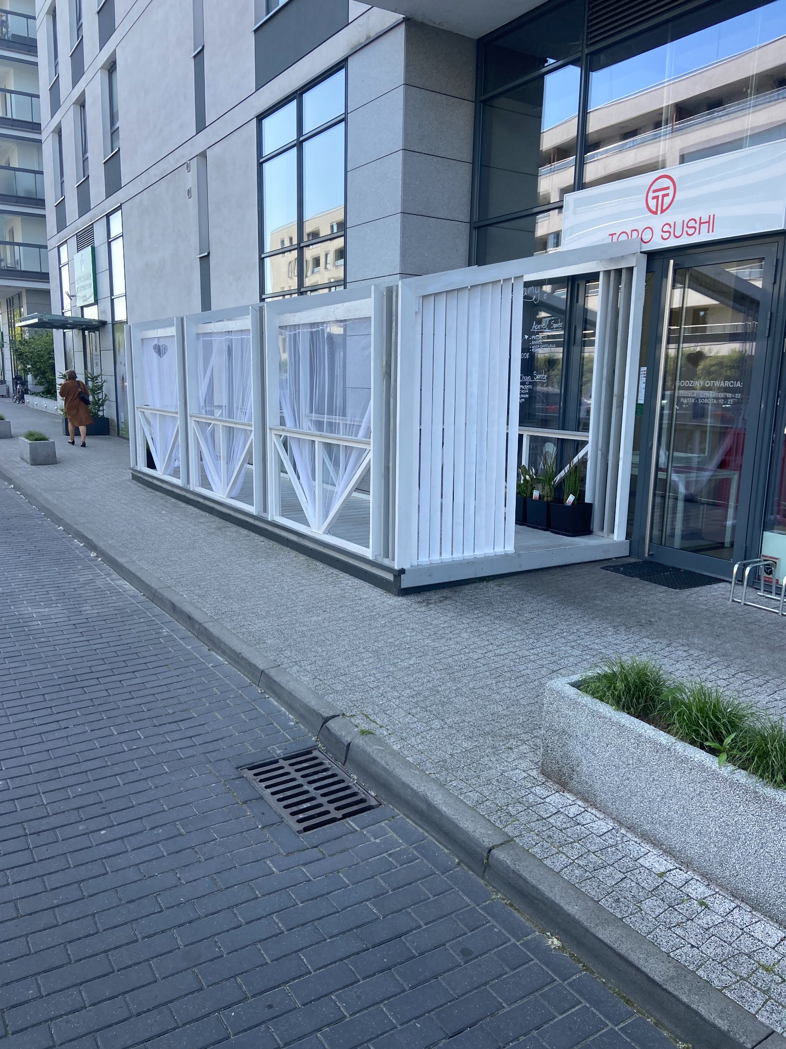 Biała, drewniana konstrukcja z zasłonami przed wejściem do restauracji Todo Sushi. Konstrukcja osłania stoliki na zewnątrz. Widoczny fragment chodnika i elewacji budynku.