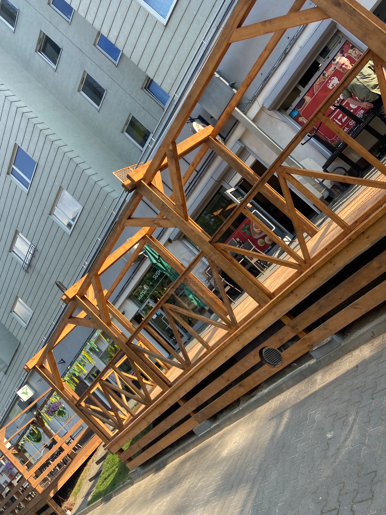 Drewniana konstrukcja tarasu przed lokalem usługowym, widoczna z perspektywy chodnika. Ażurowa balustrada z belek w ciepłym odcieniu brązu na tle budynku.