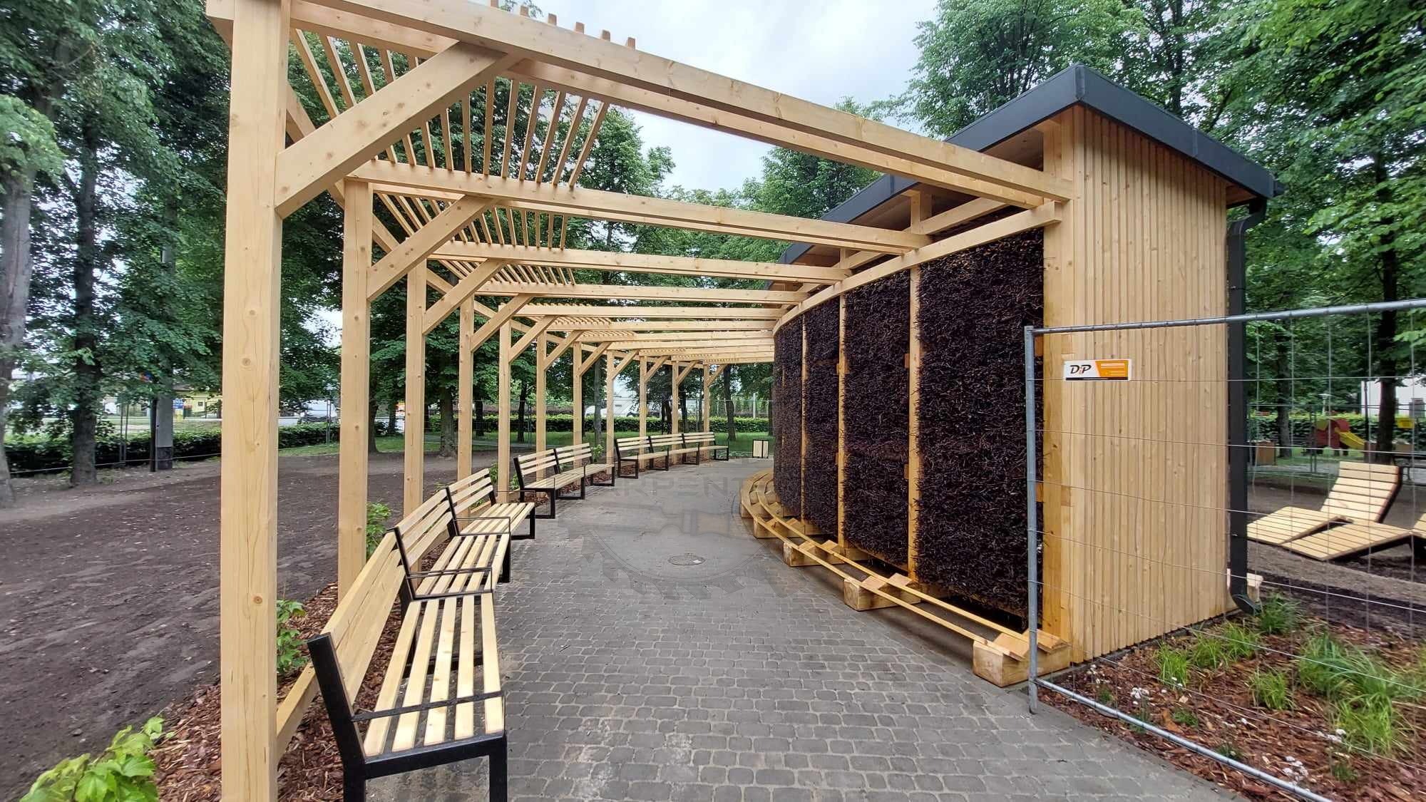 Drewniana konstrukcja tężni solankowej z ławkami w parku, wykonana z jasnego drewna, widok z perspektywy chodnika z kostki brukowej, w tle drzewa.