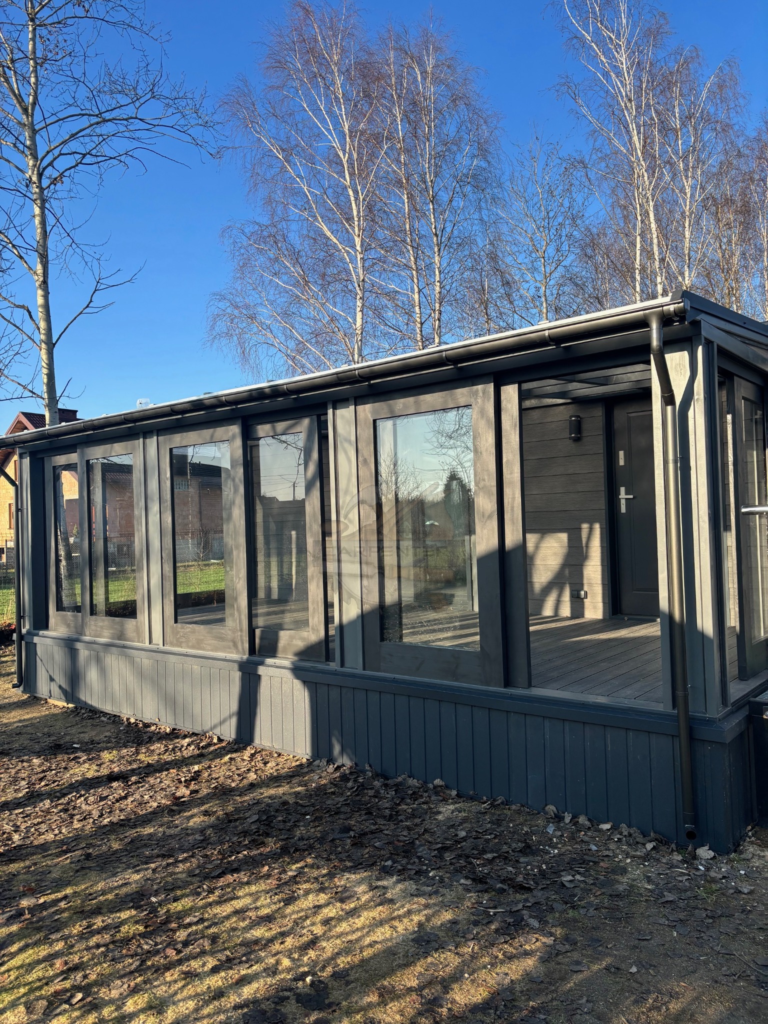 Zadaszony taras z przesuwnymi szklanymi drzwiami w kolorze grafitowym, otoczony drzewami. Widoczne refleksy w szybach i cień na ziemi. Słoneczny dzień.