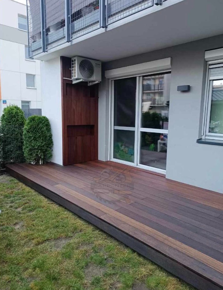 Nowoczesny balkon z drewnianą podłogą i zabudową, klimatyzator na ścianie. Widok na zewnątrz przez duże okno balkonowe. Estetyczne wykończenie.