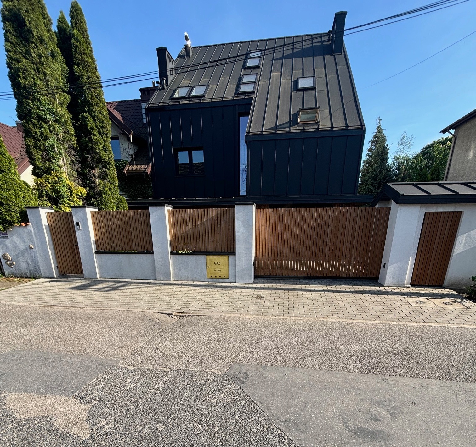 Nowoczesny dom z czarną blachą trapezową na elewacji i dachu, drewnianym ogrodzeniem z białymi słupkami. Widok z ulicy na fasadę budynku.