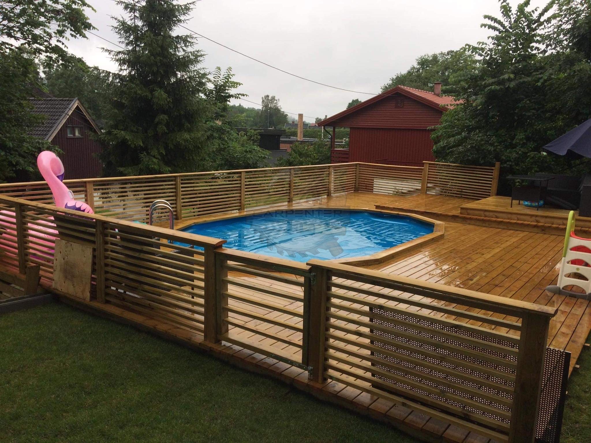 Drewniany taras z basenem, otoczony ażurowym ogrodzeniem. W tle zabudowa jednorodzinna i zieleń. Na tarasie dmuchany flaming.
