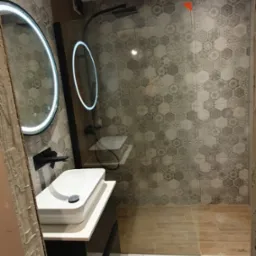 Nowoczesna łazienka z umywalką nablatową, okrągłym lustrem LED i kabiną prysznicową typu walk-in. Na ścianach heksagonalne płytki w odcieniach szarości.