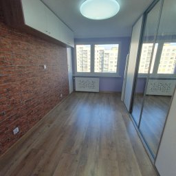 WSZYSTKO ALBO NIC NIKOLA OSIŃSKA - Wykończone wnętrze pokoju: ściana z cegły, drewniana podłoga, szafa z lustrami i okno z widokiem na budynki. Białe szafki wiszące pod sufitem.