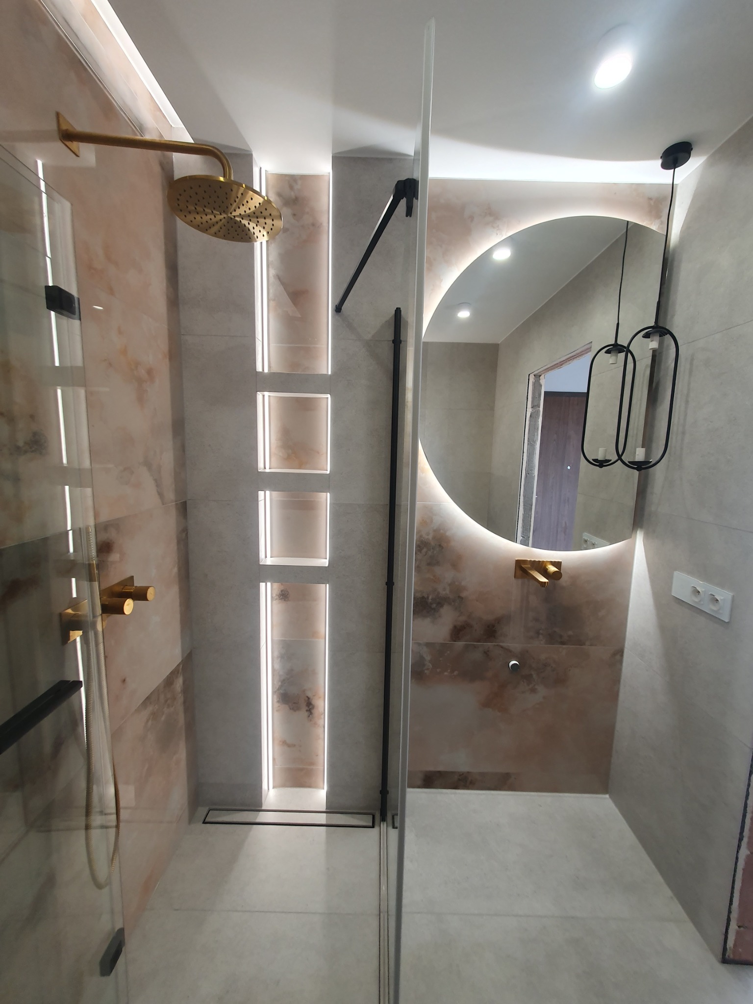Nowoczesna łazienka po remoncie: kabina prysznicowa ze złotą armaturą, okrągłe lustro z podświetleniem LED i dekoracyjne wnęki z oświetleniem.