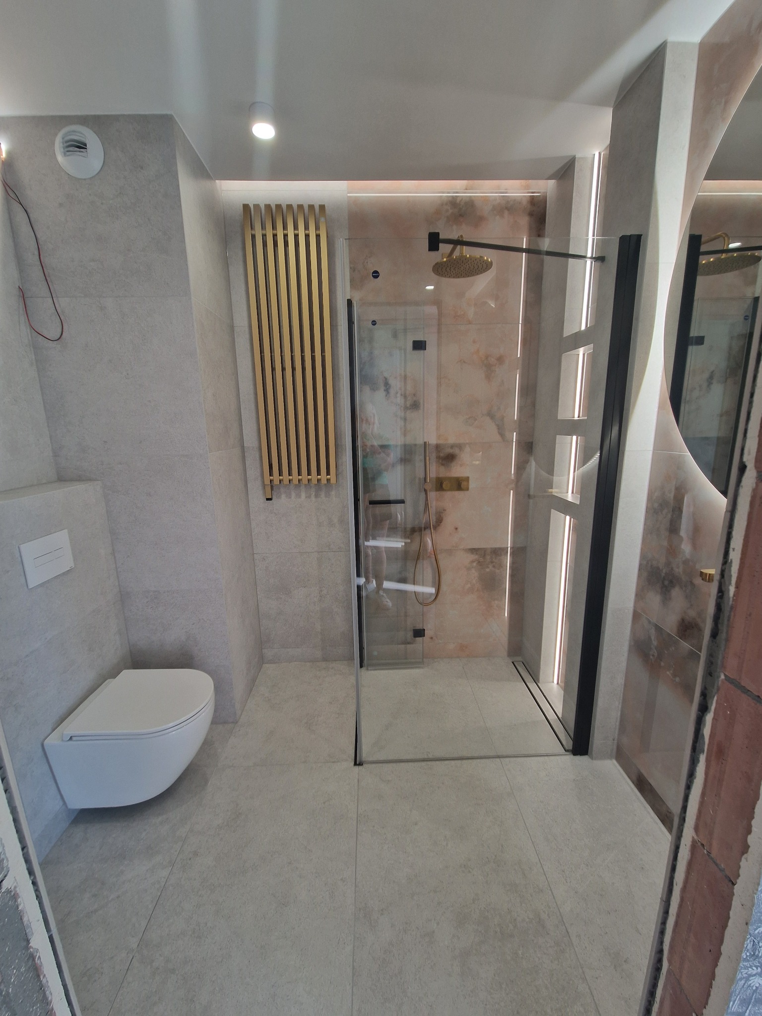 Wykończona łazienka z kabiną typu walk-in, grzejnikiem drabinkowym w kolorze złota i podwieszaną toaletą. Płytki w odcieniach szarości i beżu. Nowoczesny design.