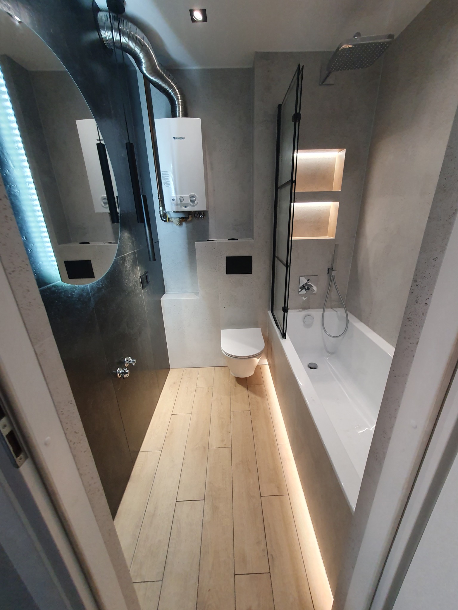 Nowoczesna łazienka w szarościach z drewnopodobną podłogą, oświetleniem LED, kabiną prysznicową typu walk-in i wiszącym piecem gazowym.