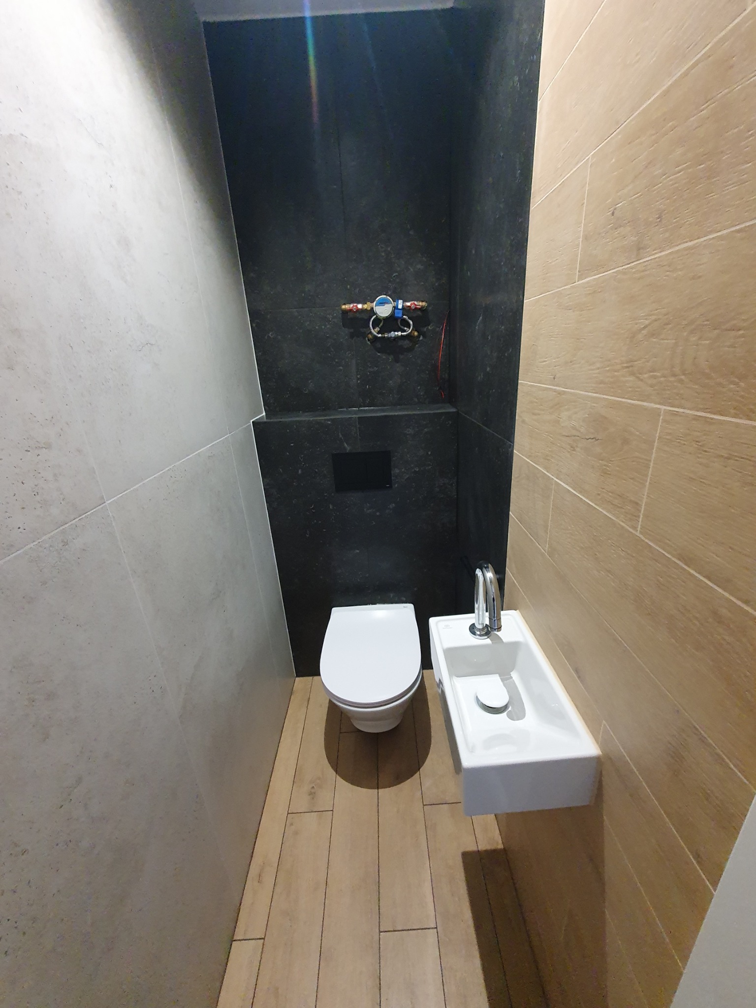 Wąska toaleta z umywalką w bloku, wykończona płytkami imitującymi drewno i kamień. Armatura zamontowana na czarnej ścianie. Widok z góry.