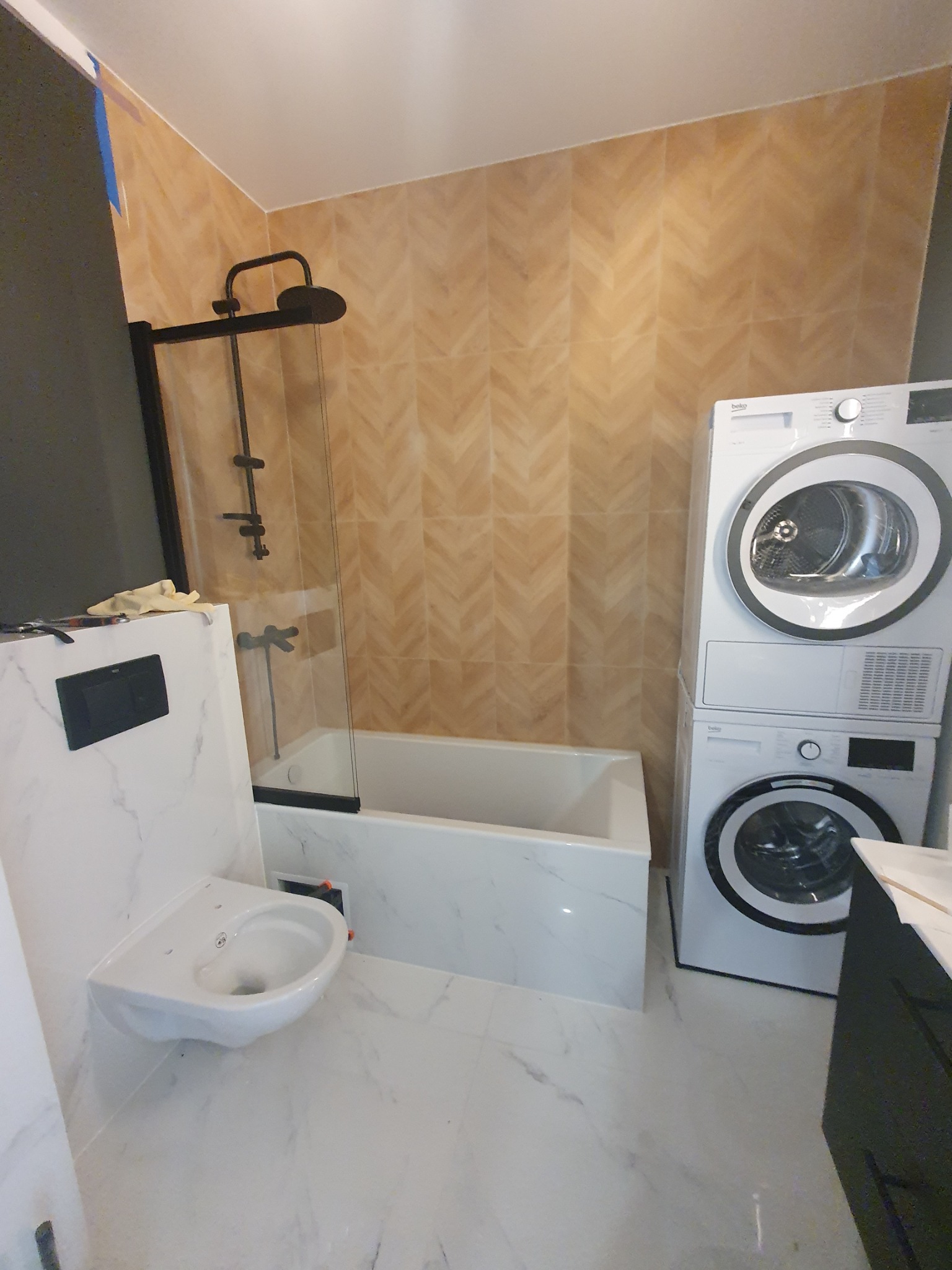 Nowoczesna łazienka z marmurową umywalką, wanną i pralko-suszarką Beko. Ściany wyłożone beżowymi płytkami w jodełkę. Czarna armatura i minimalistyczny design.