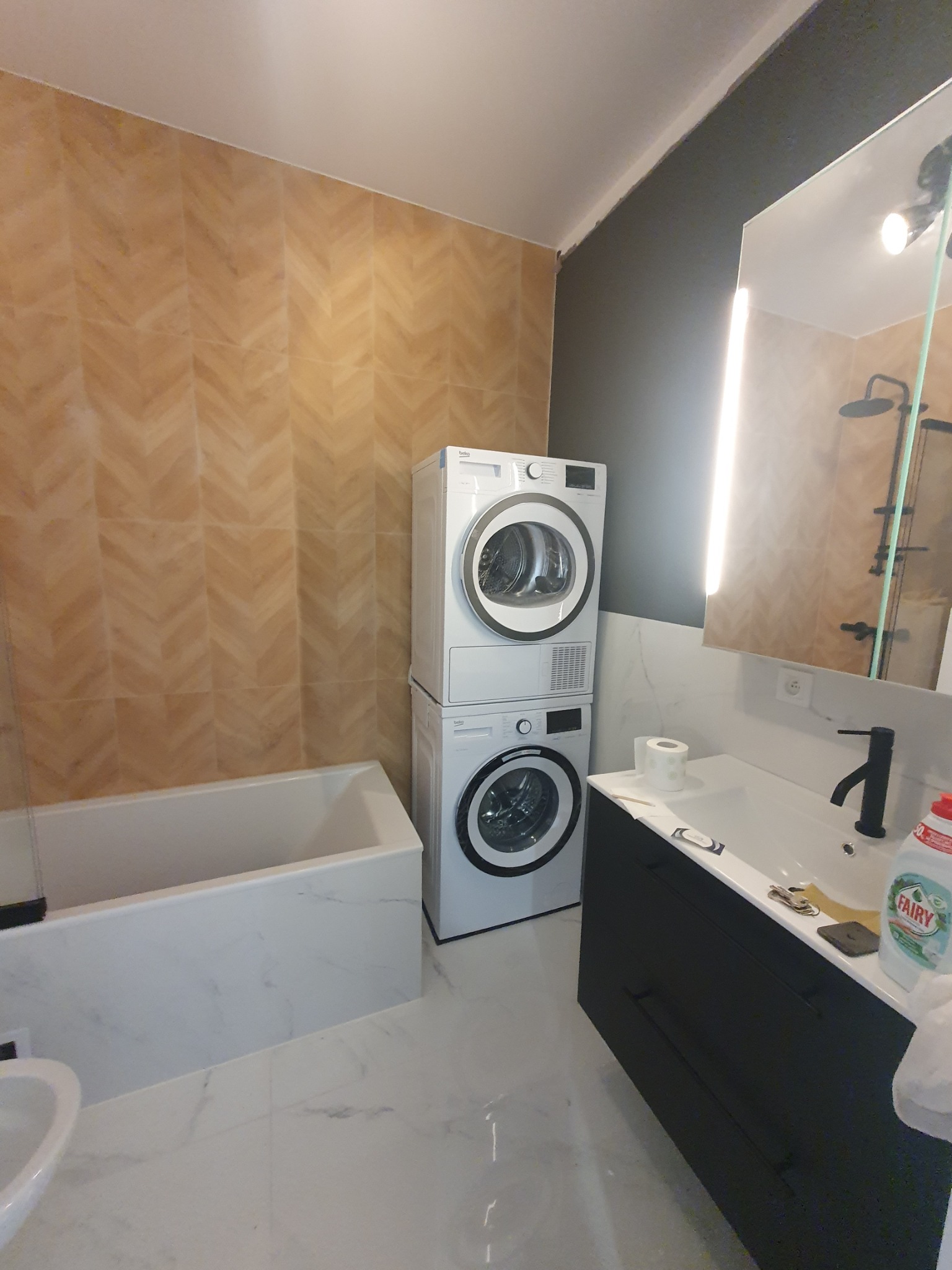 Nowoczesna łazienka po remoncie: beżowe płytki w jodełkę, biała wanna obudowana marmurem, pralka z suszarką w kolumnie i czarna szafka pod umywalką.