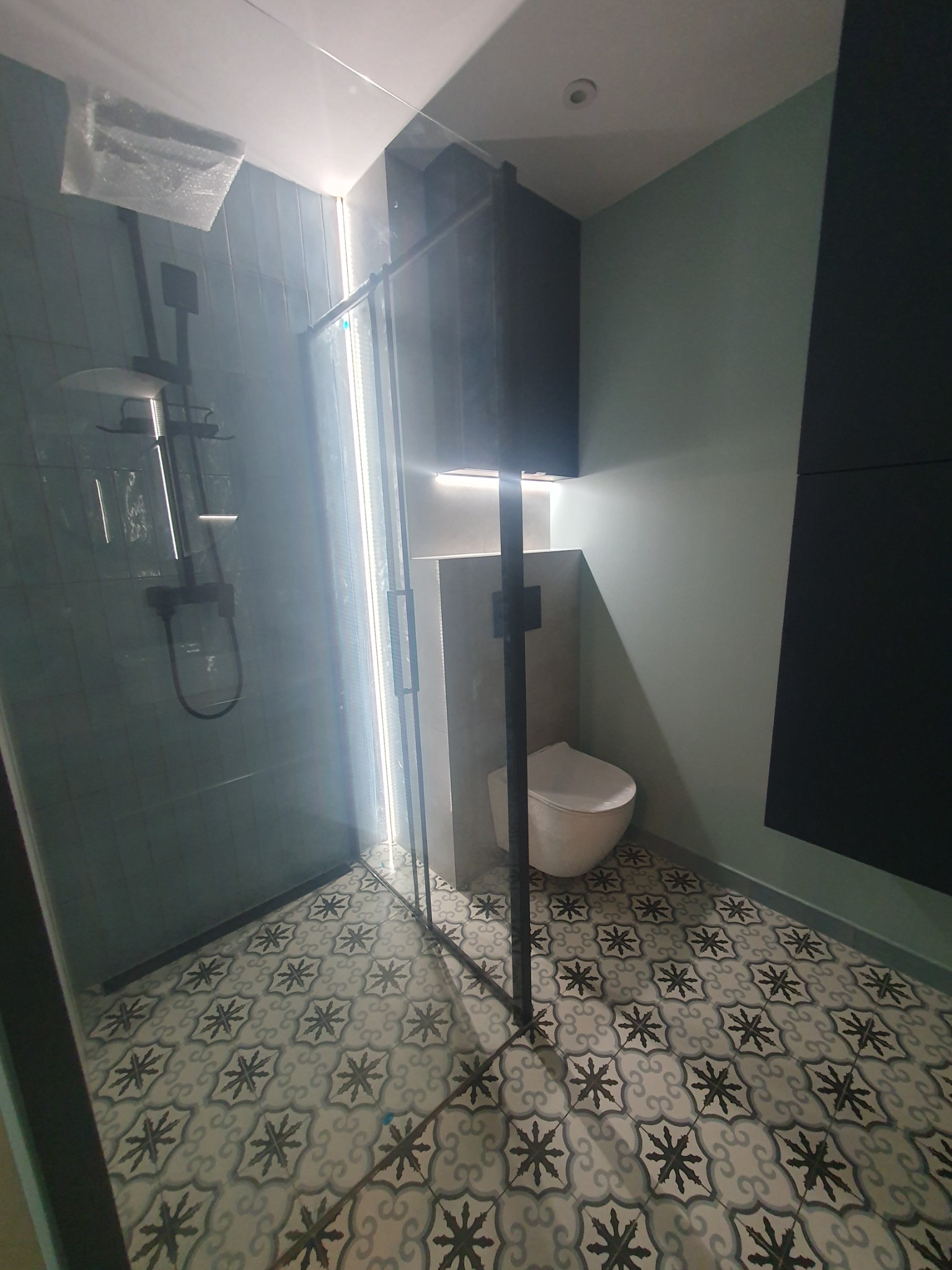 Nowoczesna łazienka po remoncie: kabina prysznicowa z czarnym zestawem, wzorzysta podłoga i toaleta podwieszana. Minimalistyczny design, świeżo po wykończeniu.