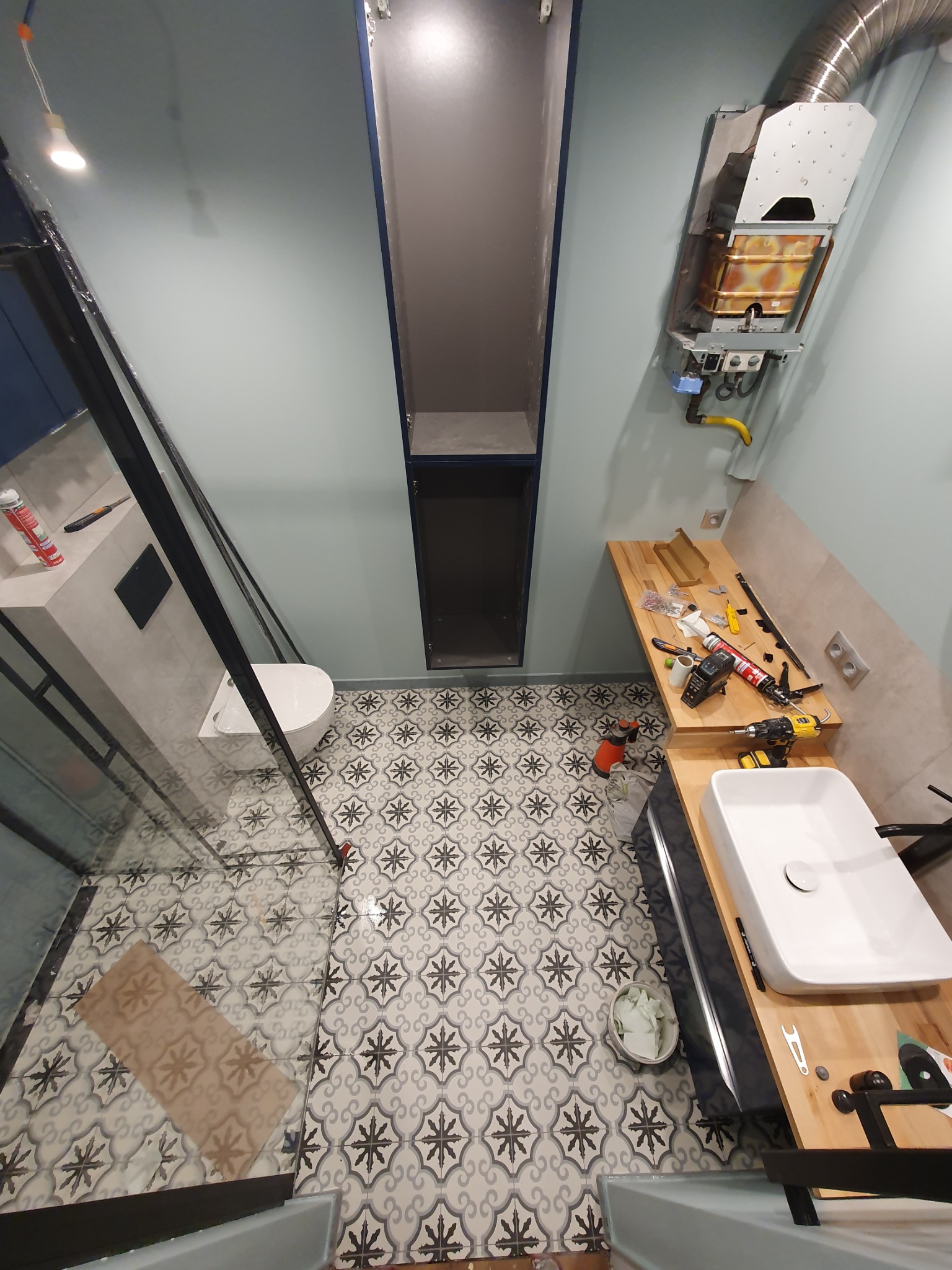 Montaż kabiny prysznicowej i umywalki w łazience z wzorzystą podłogą. Widoczne narzędzia i bojler. Perspektywa z góry.