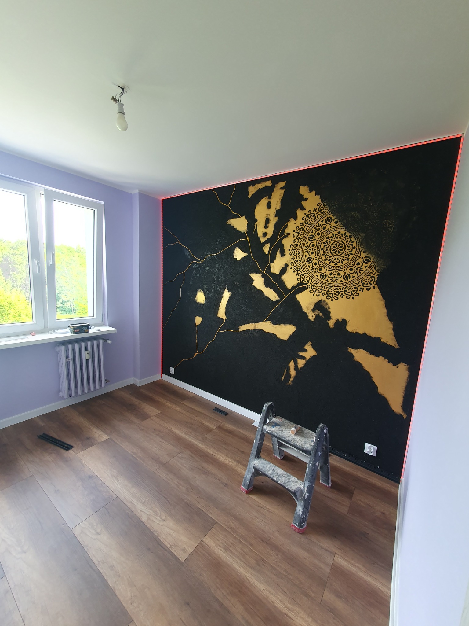 Pokój po malowaniu ścian z unikalnym wzorem w kolorze czarnym i złotym, drabina malarska stoi przy ścianie, widok na okno i grzejnik.