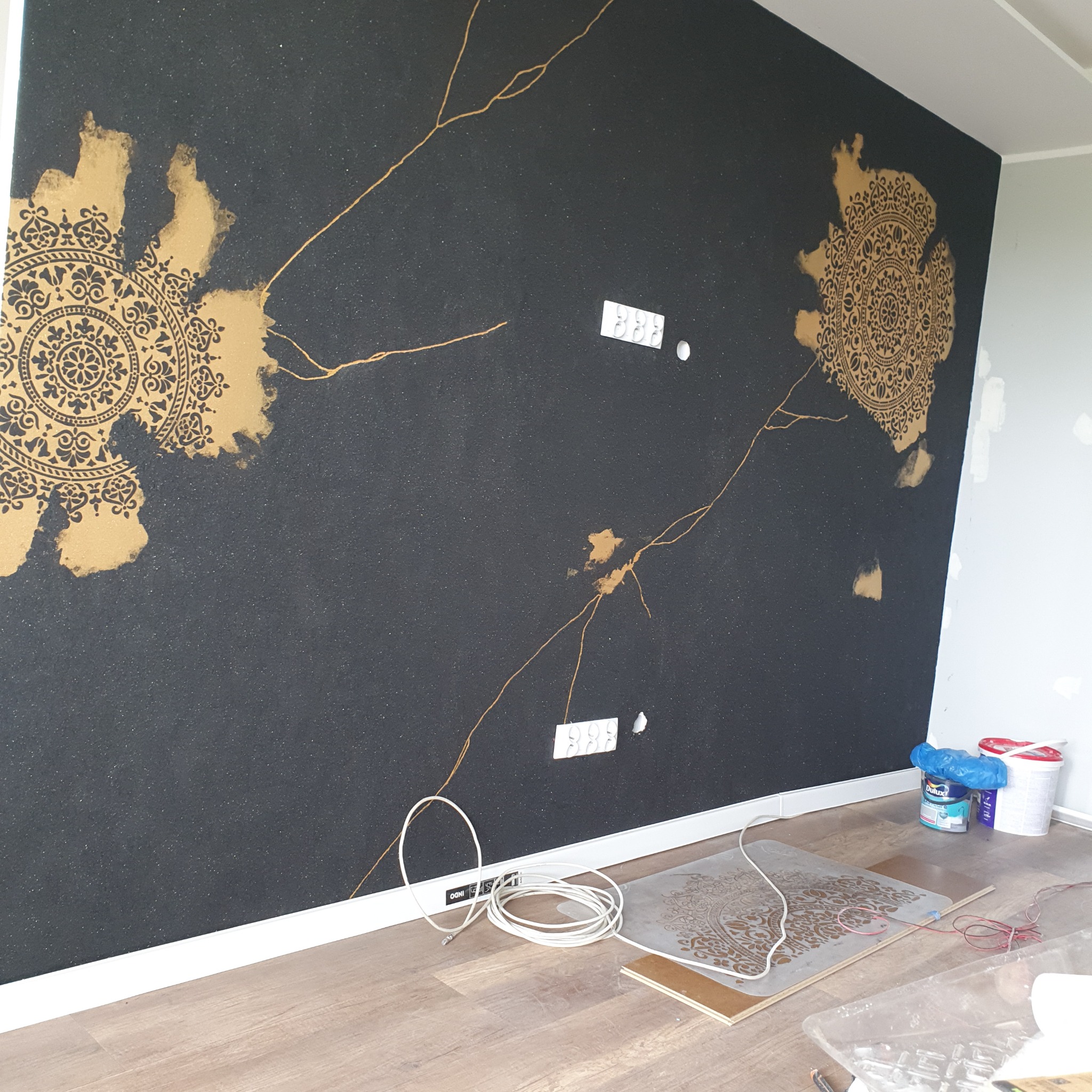 Czarna ściana z dekoracyjnymi, złotymi wzorami mandali i żyłkowaniem. Na podłodze szablony, kable i puszki farb.