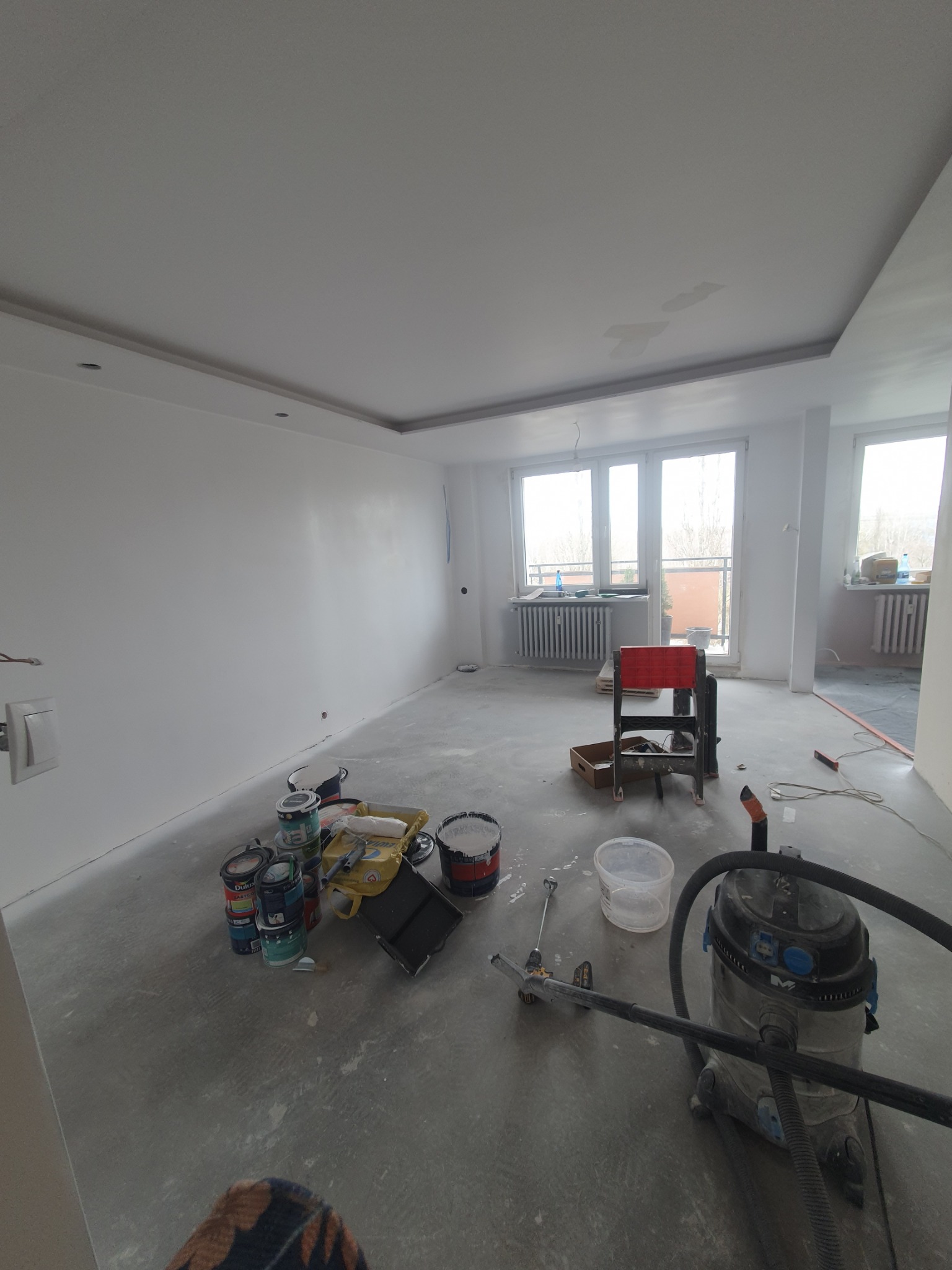 Pokój po malowaniu ścian na biało. Widoczne puszki z farbą, narzędzia malarskie i odkurzacz przemysłowy. Ściany świeżo pomalowane, z widocznymi niedoskonałościami.