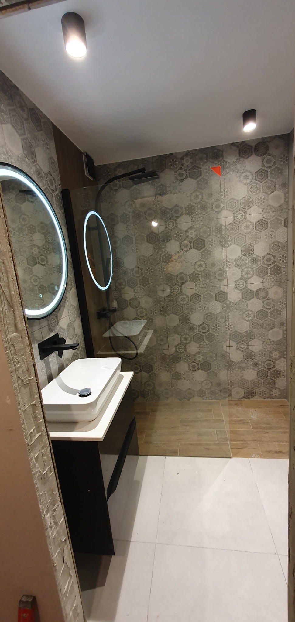 Nowoczesna łazienka z umywalką nablatową, okrągłym lustrem LED i kabiną prysznicową typu walk-in. Na ścianach heksagonalne płytki w odcieniach szarości.