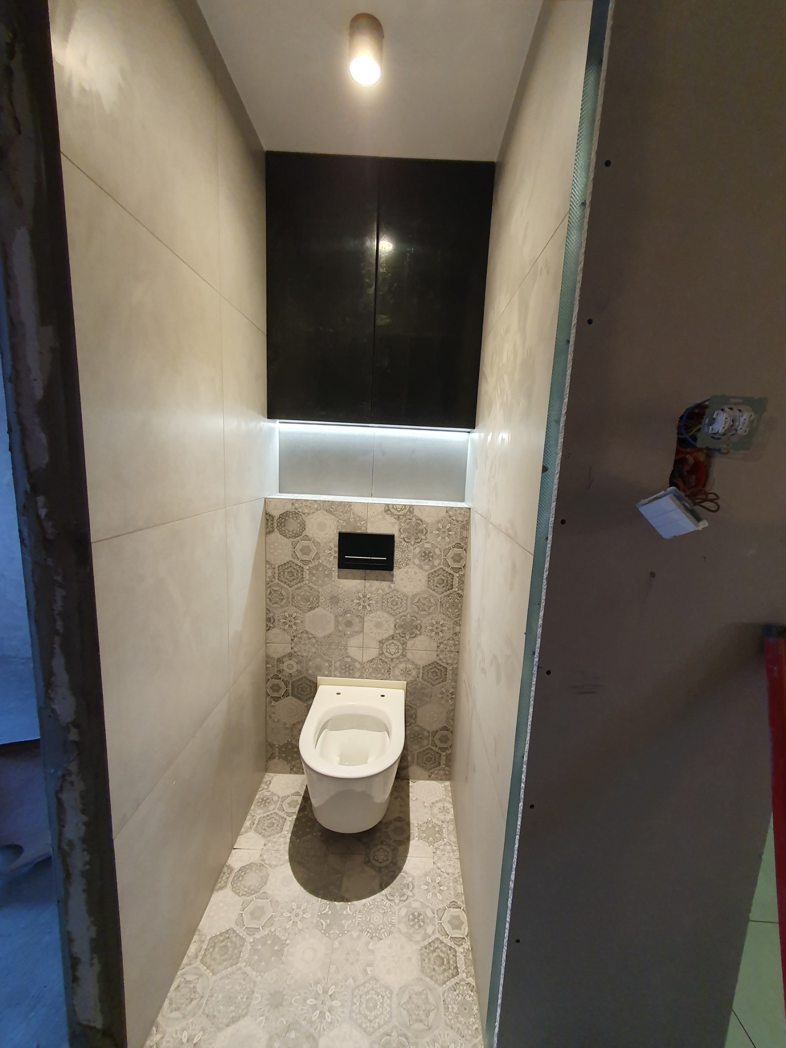 Nowoczesna toaleta w trakcie wykończenia: płytki heksagonalne, podświetlana szafka, surowa płyta gipsowa. Widoczne detale instalacji elektrycznej.