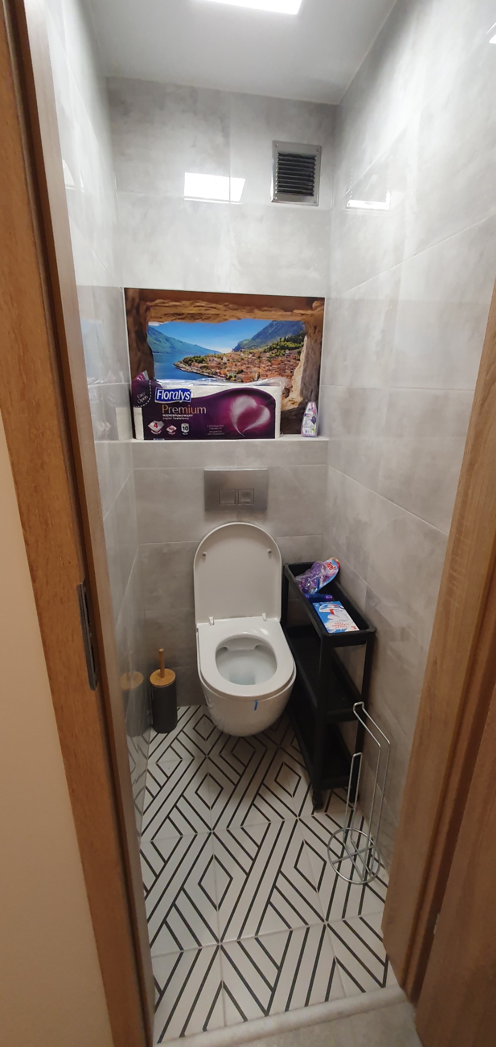 Jasna, wykończona łazienka z toaletą, wzorzystą podłogą i dekoracją ścienną imitującą widok z okna. Uwagę przykuwa czarny regał na akcesoria.