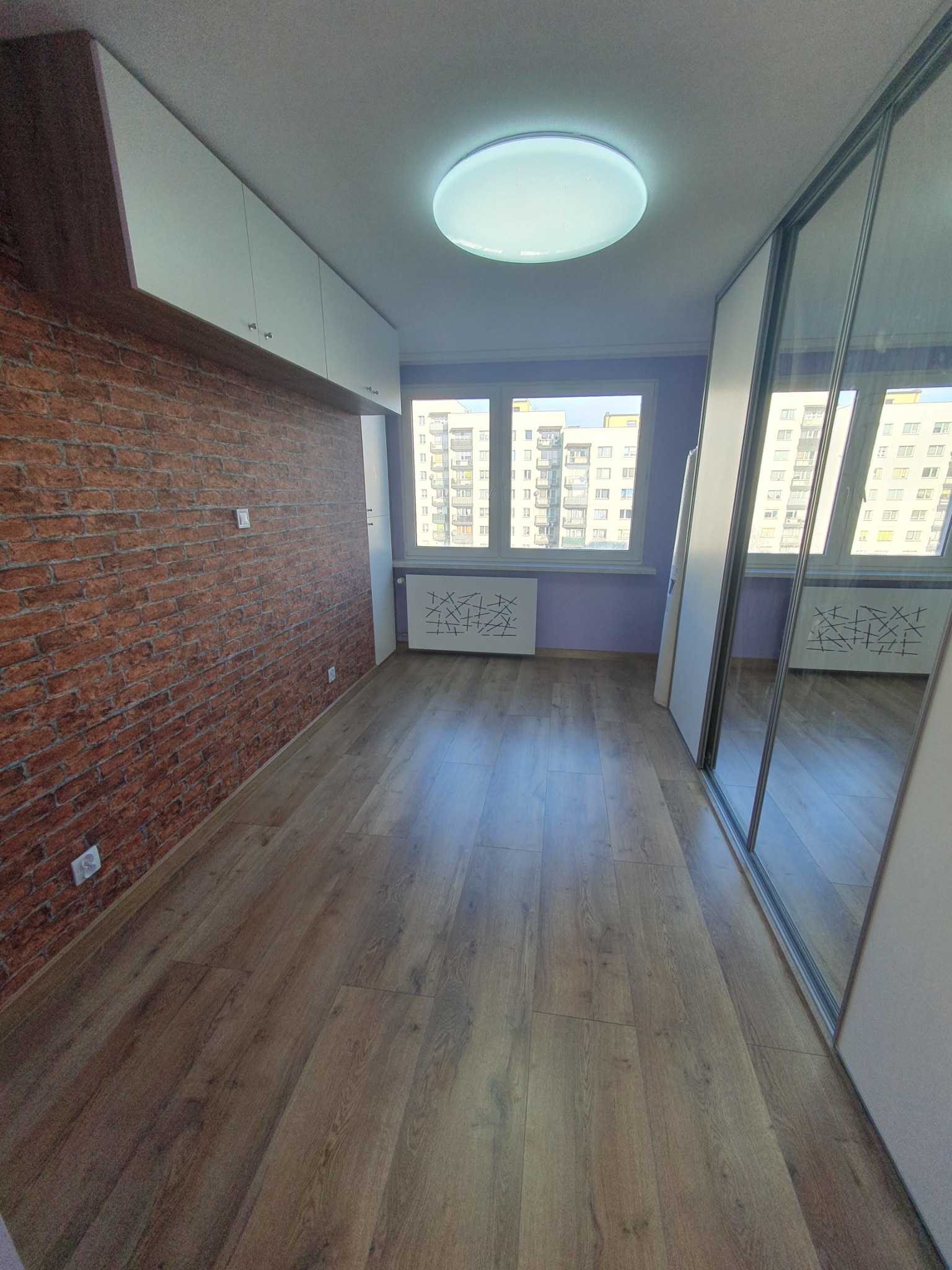 Wykończone wnętrze pokoju: ściana z cegły, drewniana podłoga, szafa z lustrami i okno z widokiem na budynki. Białe szafki wiszące pod sufitem.