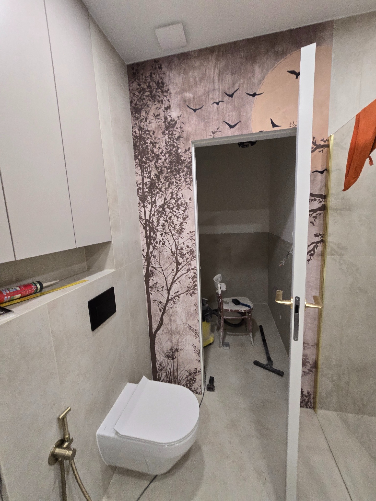 Wykończona toaleta w beżach z motywem drzewa i ptaków na ścianie. Widoczna podwieszana miska WC, szafki i bidetka. W tle pomieszczenie w trakcie prac wykończeniowych.
