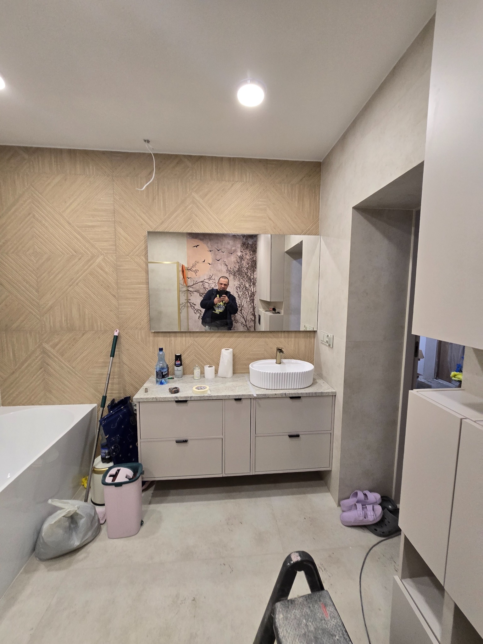 Wykończona łazienka z umywalką na szafce, lustrem i płytkami imitującymi drewno. Widoczne akcesoria i drabina malarska. Ujęcie z dołu, podkreślające przestrzeń.