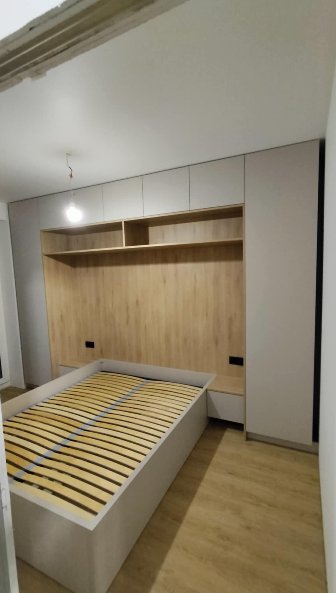 Nowoczesna szafa wnękowa z drewnianym panelem nad łóżkiem i półkami, w odcieniach szarości i drewna, z widocznym stelażem łóżka i podłogą z paneli.