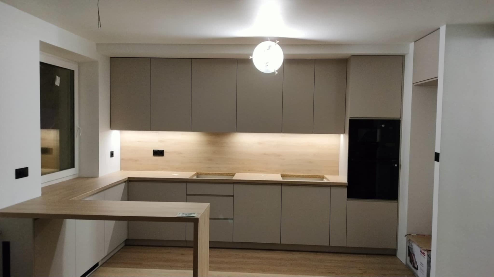 Jasna kuchnia w kształcie litery U z drewnianym blatem i oświetleniem LED pod szafkami. Widoczny czarny piekarnik i lampa sufitowa w kształcie kuli.