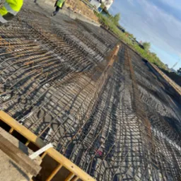 Szeroki kadr na zbrojenie fundamentów pod budynek mieszkalny, z widocznymi pracownikami w odblaskowych kamizelkach i elementami szalunków, w słoneczny dzień.