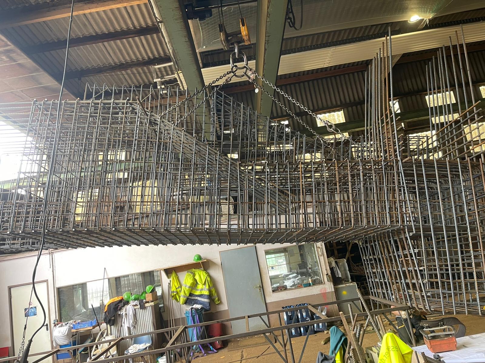 Gotowy szkielet zbrojeniowy podwieszony pod sufitem hali, przygotowany do transportu. Widoczne elementy konstrukcyjne budynku i odzież ochronna.