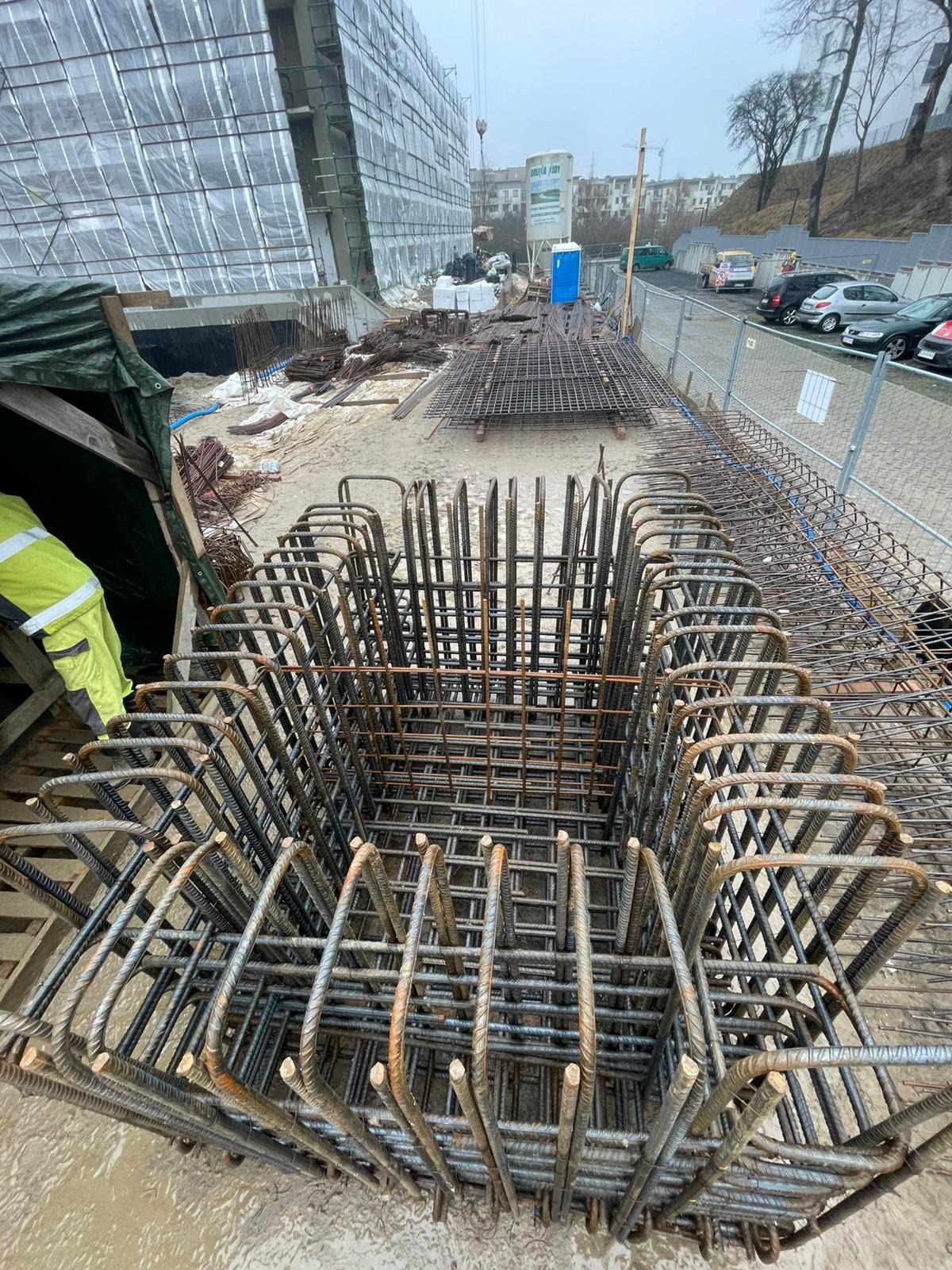 Klatka zbrojeniowa na budowie, widok z góry. Stalowe pręty tworzą szkielet konstrukcji, w tle budynek w budowie i plac budowy.