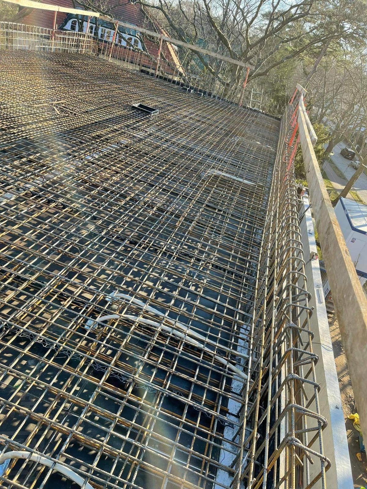 Zbrojenie stropu budynku z widocznymi przewodami instalacji elektrycznej, ujęcie z góry pod kątem, z elementami szalunku i widokiem na otoczenie.