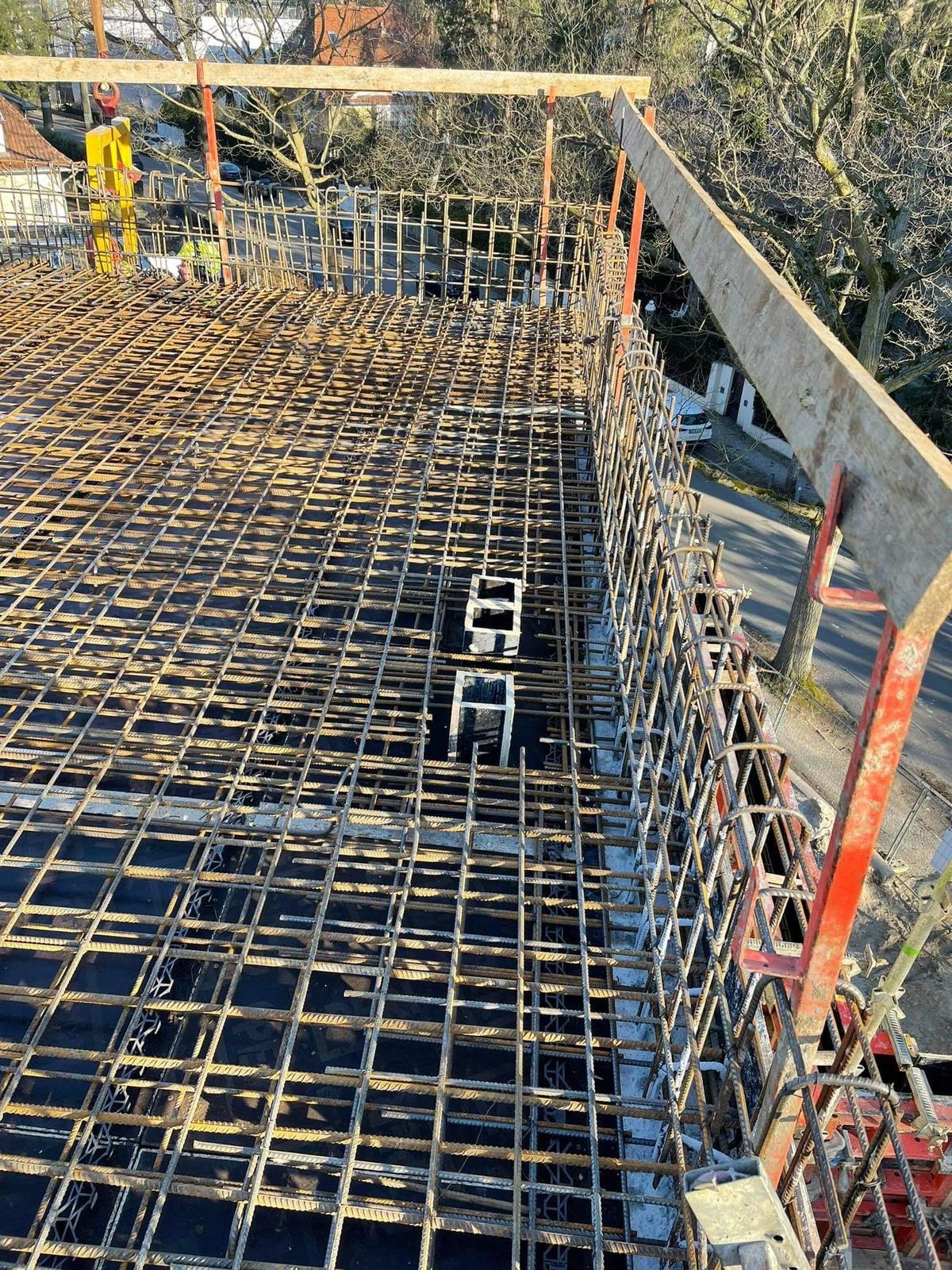 Zbrojenie stropu budynku wielorodzinnego, widok z góry. Stalowe pręty tworzą regularną siatkę. Fragment ulicy i drzewa w tle. Czerwone podpory szalunku.