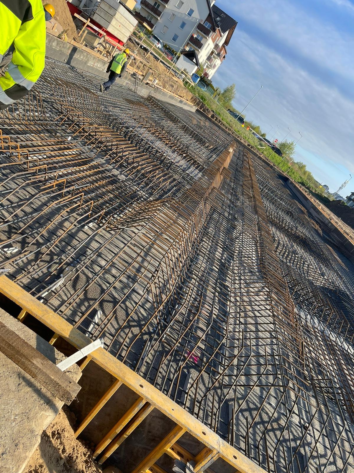 Szeroki kadr na zbrojenie fundamentów pod budynek mieszkalny, z widocznymi pracownikami w odblaskowych kamizelkach i elementami szalunków, w słoneczny dzień.