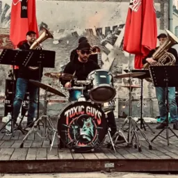 Zespół muzyczny 'Toxic Guys' na plenerowej scenie. Muzycy grają na instrumentach dętych i perkusji. W tle mur z graffiti i czerwone parasole.