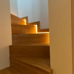 Drewniane schody z podświetleniem LED między stopniami, widok z dołu, oświetlenie ciepłe, minimalistyczne wnętrze z białymi ścianami, nowoczesny design.