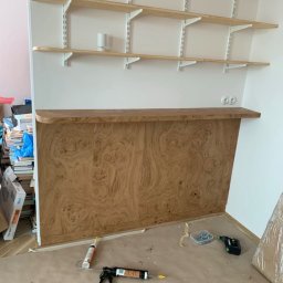 REORGET SPÓŁKA Z OGRANICZONĄ ODPOWIEDZIALNOŚCIĄ - Montaż drewnianej zabudowy pod półkami na ścianie. Widoczne narzędzia i materiały montażowe. Wykończenie z forniru o ciekawym wzorze.