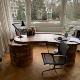 Biurko z drewna o nieregularnym kształcie, z widoczną strukturą słojów, stoi w pokoju z dużym oknem i parkietem w jodełkę. Krzesło biurowe obok.